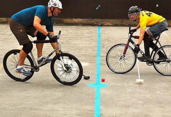 Two cyclists playing bike polo with red ball near blue center line