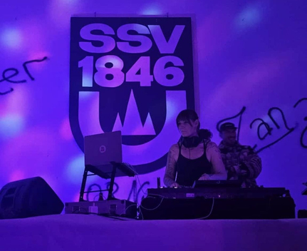 DJ an Mischpult vor SSV 1846 Banner bei Abschiedsparty in lila Licht