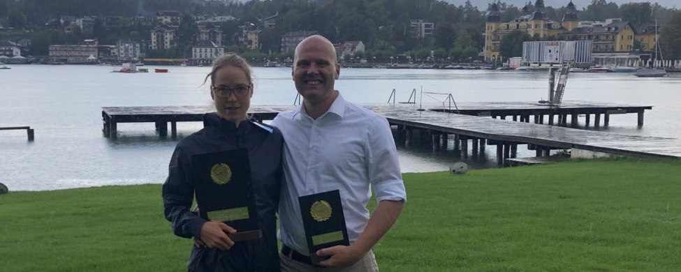 zwei Personen am Seeufer halten Trophäen vor Steg und Bergkulisse