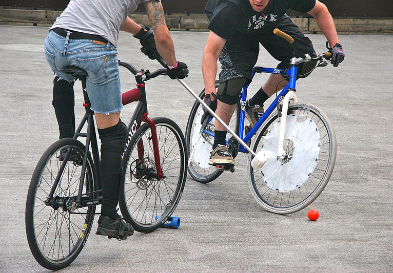 Zwei Bikepolo-Spieler auf Fahrrädern mit Schlägern und orangefarbenem Ball