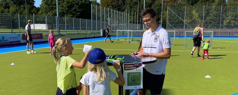 Trainer an Stehtisch mit zwei Kindern und Hockeyschlägern auf Hockeyplatz