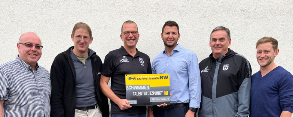 Sechs Männer halten Tafel mit Aufschrift Schwimmen Talentstützpunkt vor Wand
