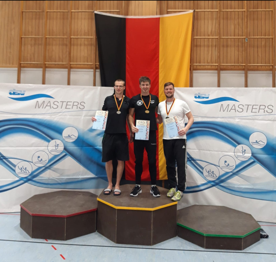Drei Schwimmer auf Podest mit Medaillen und URKUNDE vor deutscher Flagge