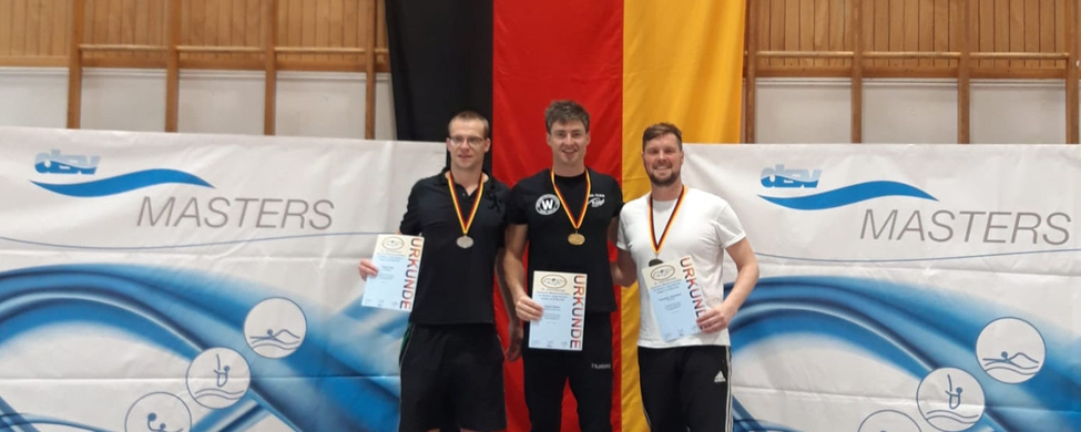 Drei Schwimmer auf Podest mit Medaillen und URKUNDE vor deutscher Flagge