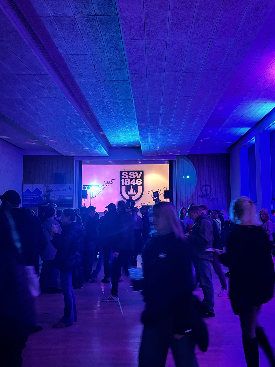 Menschen in blauviolett beleuchteter Sporthalle vor SSV 1846 Wandlogo