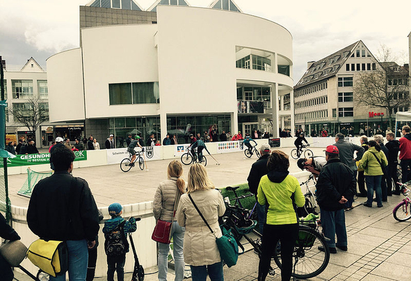 Menschen beobachten Fahrradspiel auf abgesperrtem Platz vor weißem Gebäude