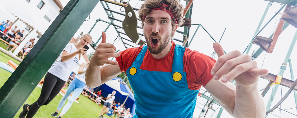 Ninja Warrior powered by Sparkasse Ulm - SSV Ulm 1846 e.V. Mann in blau-rotem Outfit gestikuliert vor Hindernisparcours