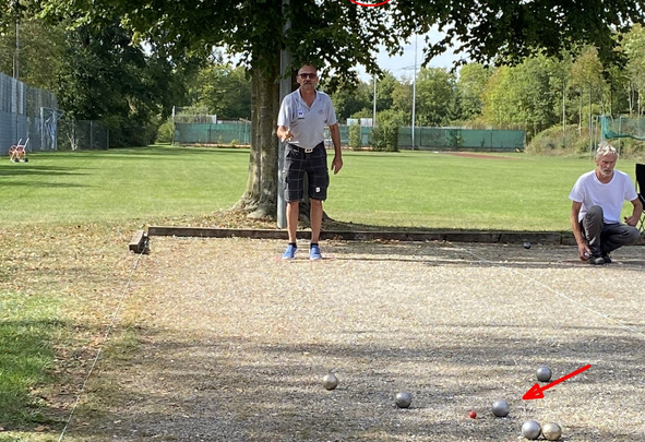 Zwei Männer spielen Boule auf Schotterbahn mit mehreren Metallkugeln und rotem Zielball