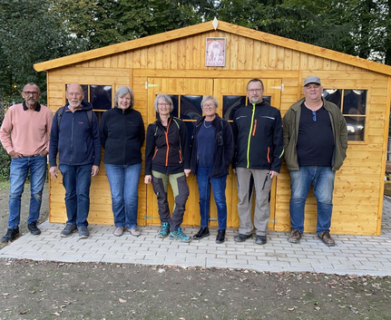 Sieben Erwachsene stehen vor einem hölzernen Gartenhaus unter Bäumen