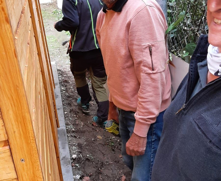 Mann mit Brille und rosa Pullover zwischen Holzwand und Zaun