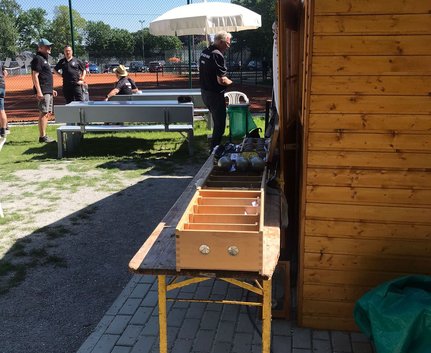 Holzhütte rechts mit Tisch Holzfach und Metallkugeln Personen an Bank vor Tennisplatz