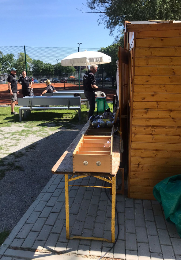 Holzhütte rechts mit Tisch Holzfach und Metallkugeln Personen an Bank vor Tennisplatz