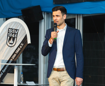 Geschäftsführer SSV Ulm 1846 e.V. Fabian Göggel führt durch den Abend. Mann im Sakko mit Mikrofon neben Banner SSV 1846 bei Sportlerehrung