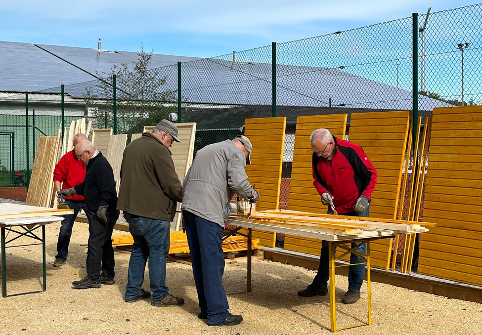 Fünf Männer streichen Holzlatten an Tischen vor gestapelten Holzwänden