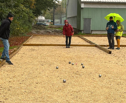 Vier Personen spielen Boule auf regennassem Schotterplatz um roten Zielball