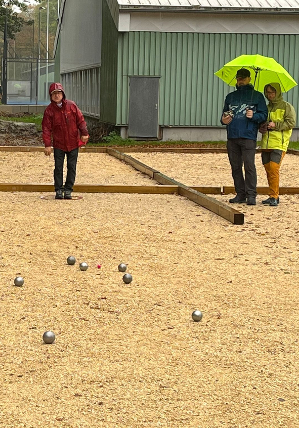 Vier Personen spielen Boule auf regennassem Schotterplatz um roten Zielball