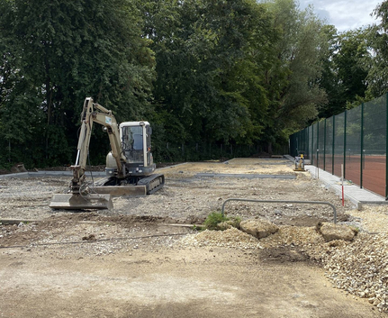 kleiner Bagger auf geschotterter Baustelle neben hohem Maschendrahtzaun