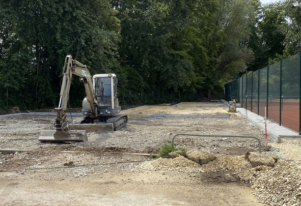 kleiner Bagger auf geschotterter Baustelle neben hohem Maschendrahtzaun