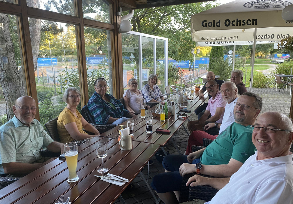 Gruppe Erwachsener an langem Holztisch unter Sonnenschirm mit Aufschrift Gold Ochsen