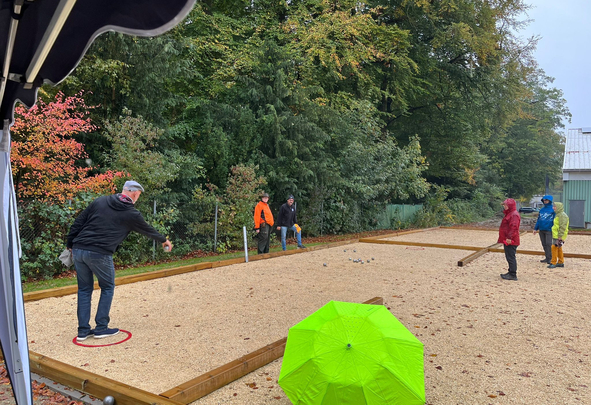 Gruppe spielt Boule auf Kiesfeld, grüner Regenschirm im Vordergrund