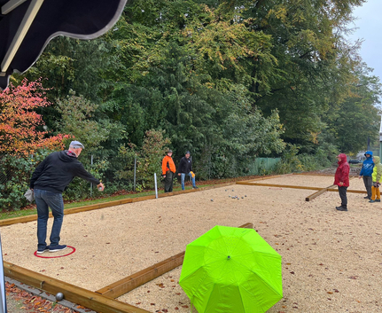 Gruppe spielt Boule auf Kiesfeld, grüner Regenschirm im Vordergrund