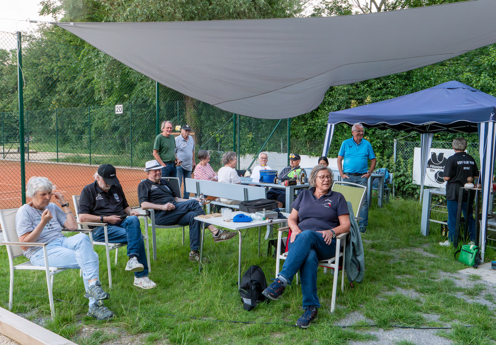 Mehrere Personen sitzen unter Sonnensegel neben Tennisplatz
