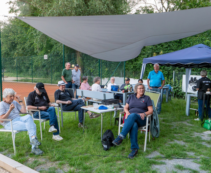 Mehrere Personen sitzen unter Sonnensegel neben Tennisplatz
