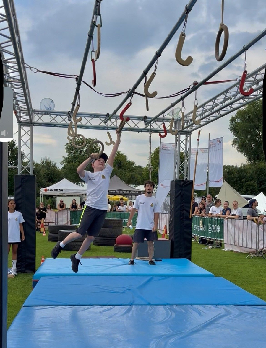 Teilnehmer springt zu hängenden Ringen im Ninja-Parcours vor Zuschauern