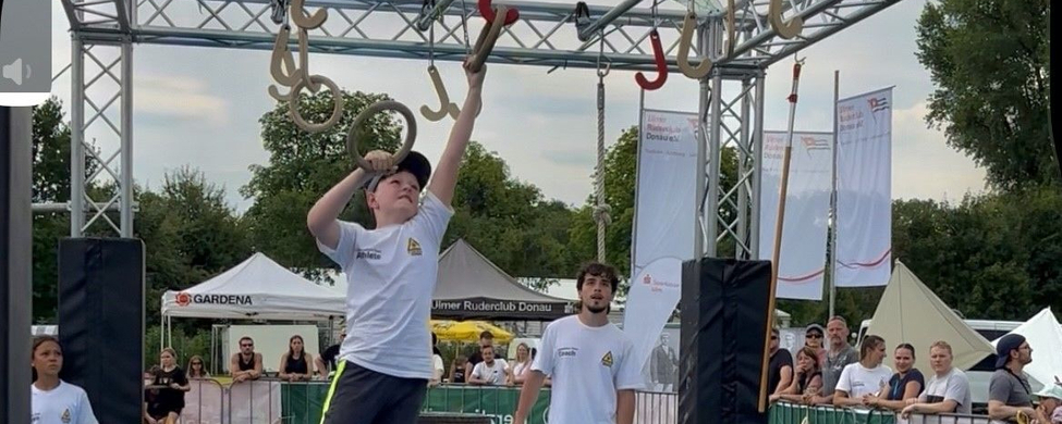 Teilnehmer springt zu hängenden Ringen im Ninja-Parcours vor Zuschauern