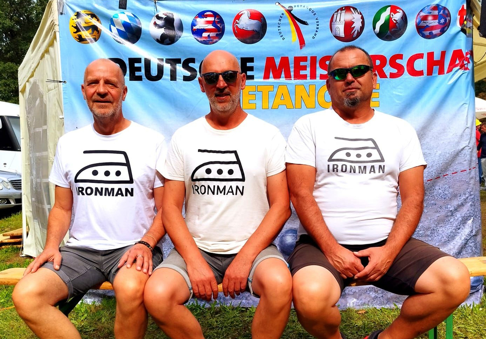 Drei Männer sitzen auf Bank in weißen T‑Shirts mit Schriftzug IRONMAN