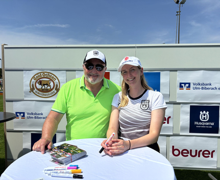 Zwei lächelnde Personen signieren Autogrammkarten vor Sponsorwand