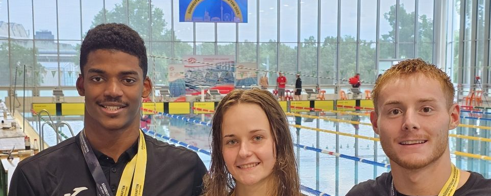 Drei Schwimmer mit Medaillen an gelben Bändern vor Wettkampfbecken