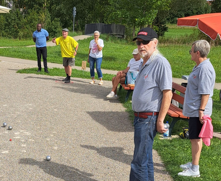 Sechs Personen spielen Boule auf Kiesfläche im Park