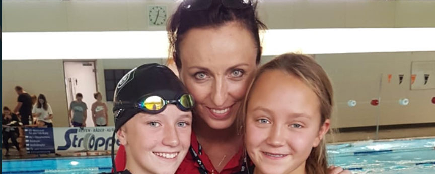 Schwimmtrainerin umarmt zwei junge Schwimmerinnen am Hallenbad