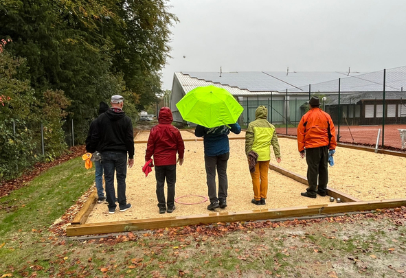 Fünf Personen auf Bouleplatz, eine mit grünem Regenschirm