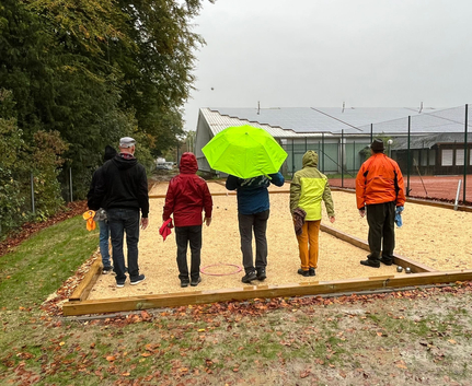 Fünf Personen auf Bouleplatz, eine mit grünem Regenschirm