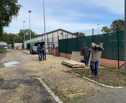 Drei Personen neben Tennisplatz, eine trägt Holzbalken