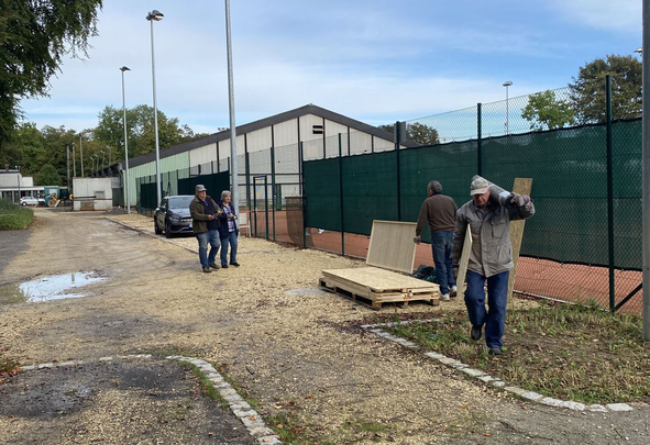 Drei Personen neben Tennisplatz, eine trägt Holzbalken