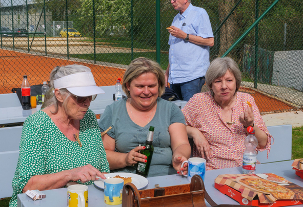 Drei Frauen sitzen an Picknicktisch neben Tennisplatz, essen Pizza und trinken