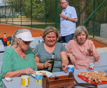 Drei Frauen sitzen an Picknicktisch neben Tennisplatz, essen Pizza und trinken