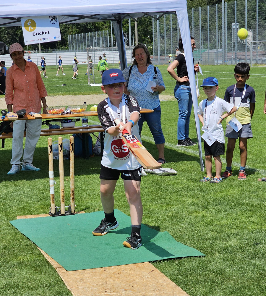 Kind schlägt mit Cricket-Schläger vor Cricket-Stand beim Schwörsporttag
