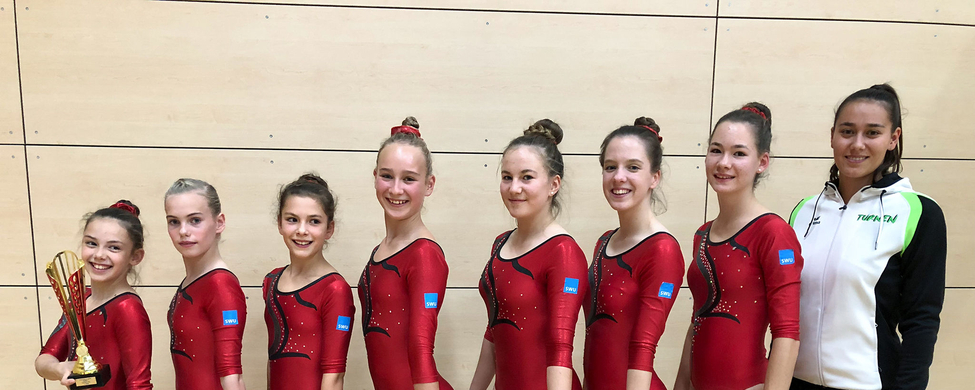 Kunstturnerinnen in roten Trikots stehen mit Pokal und Trainerin