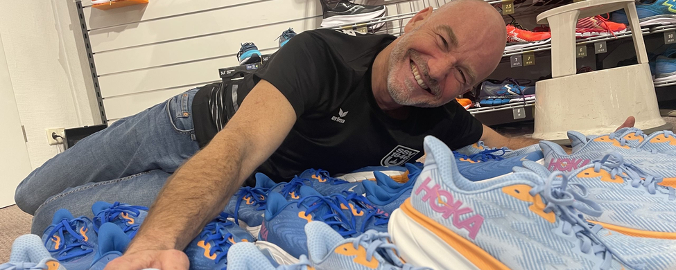 Lächelnder Mann zwischen vielen hellblauen Hoka-Laufschuhen