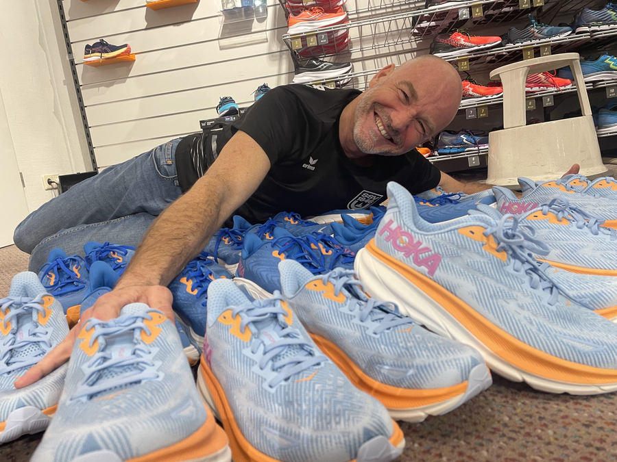 Lächelnder Mann zwischen vielen hellblauen Hoka-Laufschuhen