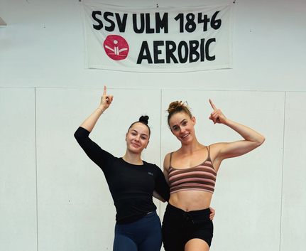 Zwei Athletinnen in Trainingskleidung zeigen auf Banner SSV ULM 1846 AEROBIC