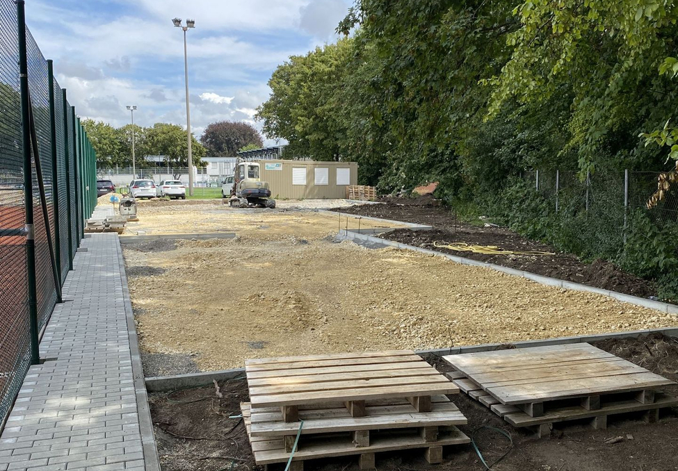 Baustelle mit gestapelten Paletten, kleinem Bagger, gepflastertem Gehweg und Bäumen