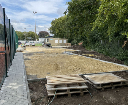 Baustelle mit gestapelten Paletten, kleinem Bagger, gepflastertem Gehweg und Bäumen