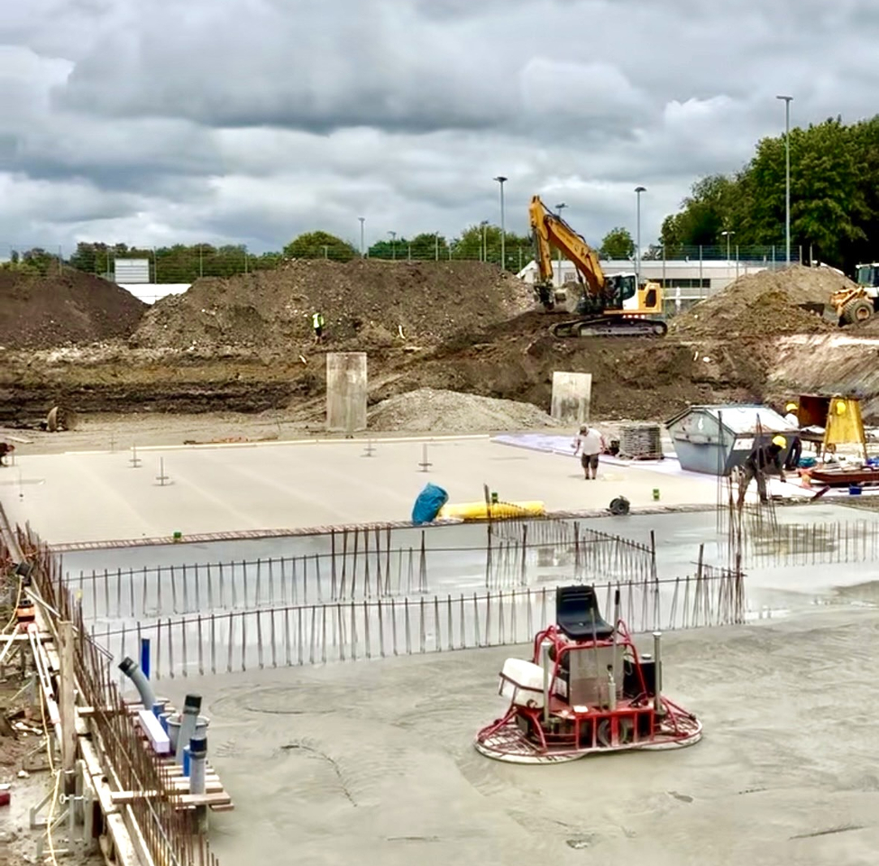 Übersicht der Baustelle Beurer Sportpark. Die Bodenplatte wird betoniert, im Hintergrund sind noch Erdmengen zu sehen und ein Bagger.