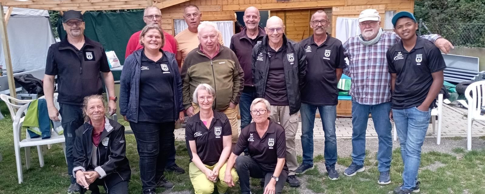 Dreizehn Boule-Spieler und Spielerinnen in Vereinskleidung vor Holzhütte