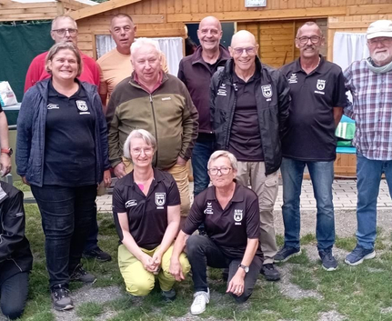 Dreizehn Boule-Spieler und Spielerinnen in Vereinskleidung vor Holzhütte